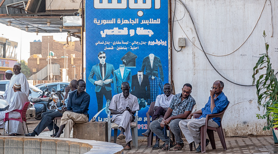 Khartoum, Sudan, February 5, 2019: Locals sitting on plastic chairs in the centre of the capital in front of a poster advertising western formal clothing