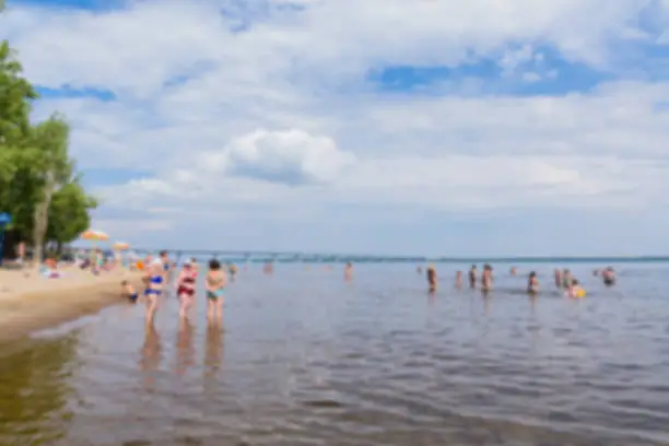 Abstract blurred image. Defocused lens, bokeh. People sunbathe and swim in the river on a sandy beach. On a Sunny summer day. Road bridge on the horizon.