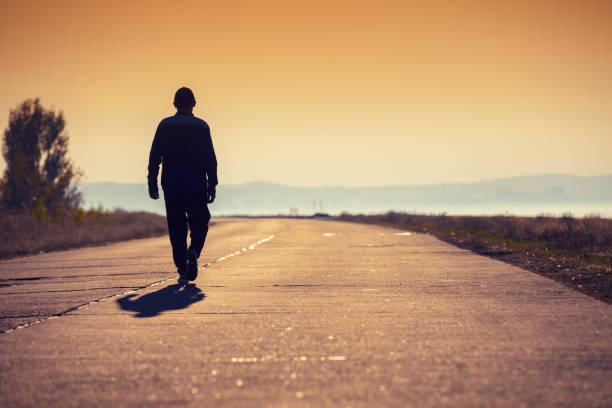 un uomo cammina lungo una strada di cemento lungo il mare in un tramonto dorato e guarda la montagna - solo un uomo foto e immagini stock