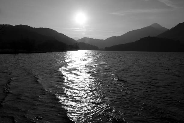 Black and white view across a lake to silhouetted mountains with setting sun View across a lake in Snowdonia with setting sun reflecting off the water. Mountains in silhouette in the background. llyn gwynant stock pictures, royalty-free photos & images