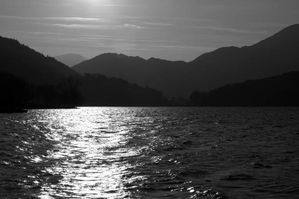 Black and white view across a lake to silhouetted mountains View across a lake in Snowdonia with setting sun reflecting off the water. Mountains in silhouette in the background. llyn gwynant stock pictures, royalty-free photos & images