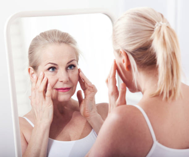 middle aged woman looking at wrinkles in mirror. plastic surgery and collagen injections. makeup. macro face. selective focus - women beauty innocence make up imagens e fotografias de stock