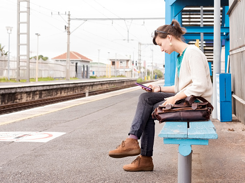 Shot of a young woman sitting on a bench at train station and using mobile phone while waiting for the train