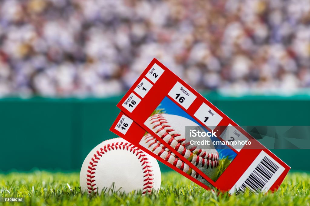 A pair of ticket stubs sitting on a baseball in the grass of a stadium A low angle view of a pair of ticket stubs to a baseball game, they are leaning on a baseball that is sitting in the outfield grass of a stadium with a green wall and crowd in the background. Ticket Stock Photo