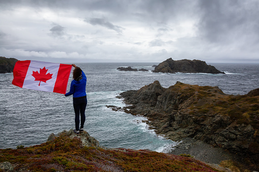 Adventurous woman holding a Canadian Flag on a Rocky Atlantic Ocean Coast during a cloudy day. Taken in Sleepy Cove, Crow Head, Twillingate, Newfoundland, Canada.