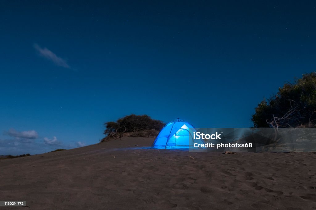 Camping on the Bani Dunes at Night Descriptive scene of the night sky with the Milky Way during the eclipse of the moon on January 21, 2019, where the Orion, Pleiades, Can Major and thousands of stars in very dark skies are clearly seen on the sand dunes of Bani in the Dominican Republic. A blue tent enhances the interest of the composition where all the elements draw attention to venturing into adventure tourism Adventure Stock Photo