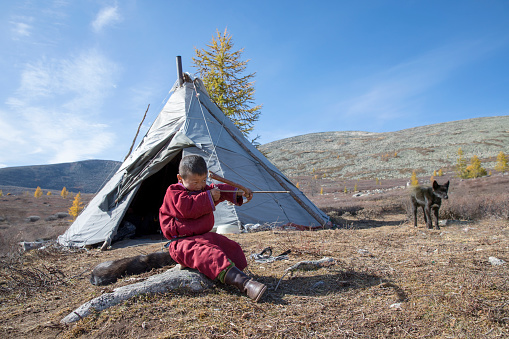 tsaatan boy in a landscape of northern mongolia practicing to shoot arrows with his home yurt at the background