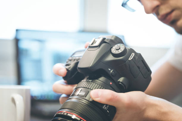 Photographer holds a reflex camera with telephoto lens in his hand. Table and laptop in the blurry background Photographer is holding a professional camera with telephoto lens in his hand, laptop in the blurry background slr camera stock pictures, royalty-free photos & images
