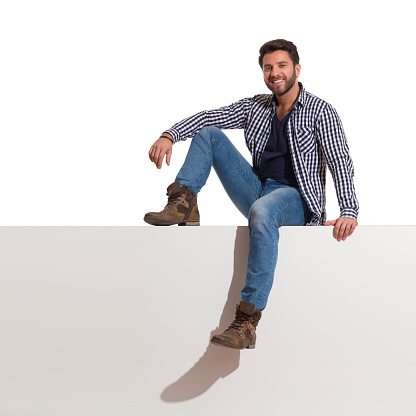 Young man in boots, jeans and unbuttoned lumberjack shirt is sitting relaxed on a top, smiling and looking at camera. Full length studio shot isolated on white.