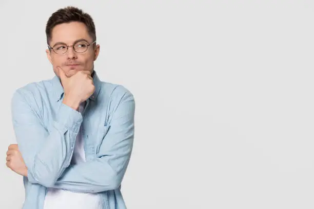 Thoughtful suspicious young man looking aside at copy space feeling skeptic doubtful, distrustful sly cunning guy thinking holding hand on chin isolated on grey white studio background