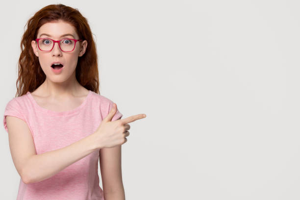 istock Amazed redhead girl student in pink glasses pointing at copyspace 1130250149