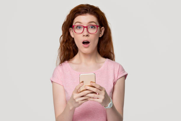 istock Shocked surprised redhead girl holding cell phone looking at camera 1130250156
