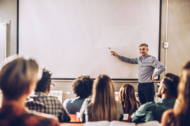 Happy professor teaching a lecture on visual screen in the classroom. Happy male teacher giving a lecture to large group of students on visual screen in the classroom. Copy space. teacher projector stock pictures, royalty-free photos & images