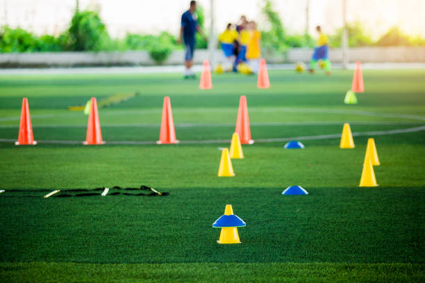 Cone markers is soccer training equipment on green artificial turf with blurry kid players training background. Material for training class of football academy