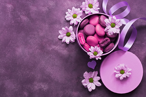 Purple and pink macaroons in a gift box on a beautiful purple background decorated with flowers. Top view, copy space.