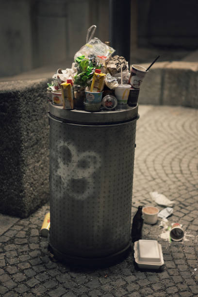 Saturday evening in Munich after a sunny day: An overflowing metallic trashcan bears witness to a successful day for the surrounding coffeeshops and supermarkets at Münchner Freiheit, in Munich's Schwabing district. The abundant garbage offers the only pops of color in an otherwise gray environment.