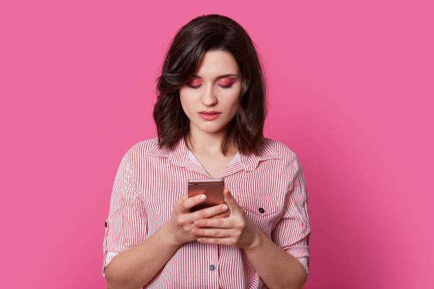 istock Brown haired girl student thinks, what happened with her touch screen phone and hung head with beautifully painted eyes. Young unique woman stands in center on isolated light pink background. 1130292414