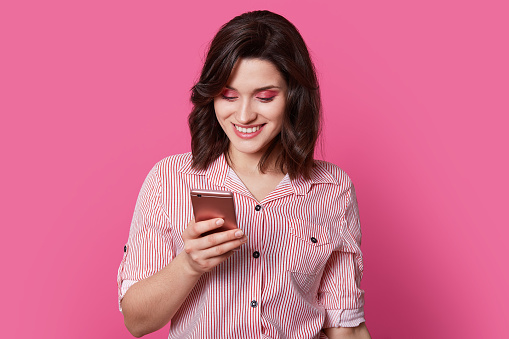 Ð¡harming, beloved female person looks into smartphone with smile in middle of portrait photography on isolated pink background. Her brown curls look great on face where bright makeup is located.