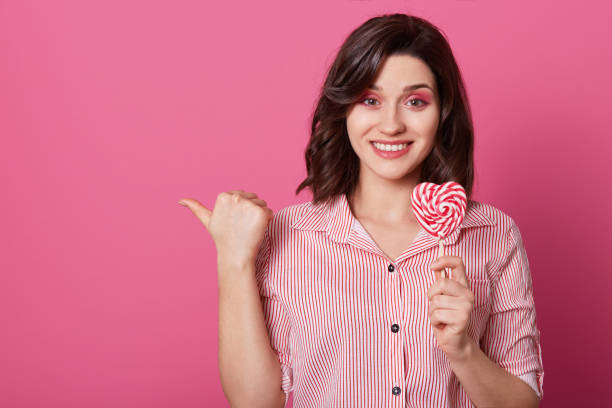 istock Horizontal shot of attractive smiling woman with pleased expression, points with thumb, holds candy in form of heart, isolated over pink background, shows place for your advertising content. 1130292467