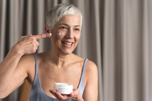 Cheerful beautiful mature woman holding jar of cosmetic product and applying cream on her face in cosy home, Beauty and cosmetic routine concept
