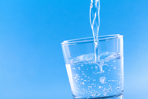 Drinking water pouring into glass against blue background closeup.