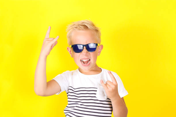 Cute blond boy wearing sunglasses on yellow background showing Horns gesture