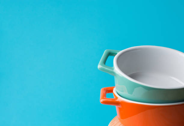 Stack of orange and turquoise ceramic cocottes on white wood table blue wall background. Cooking baking cookware concept. Scandinavian kitchen interior. Workshop poster template