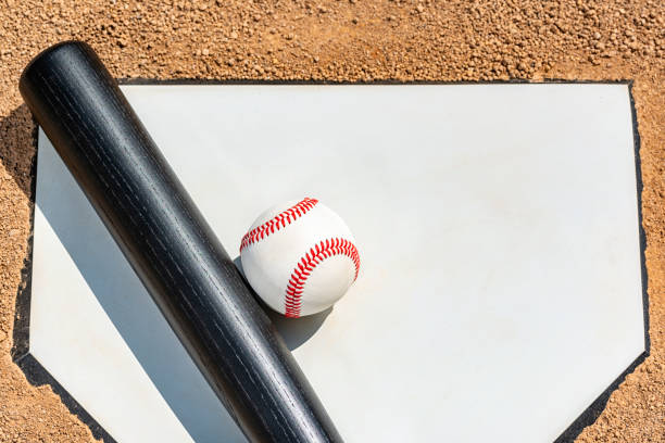 Looking down on a new baseball and a wooden bat painted black siting on home plate with hints of the white lines of the batter's box.
