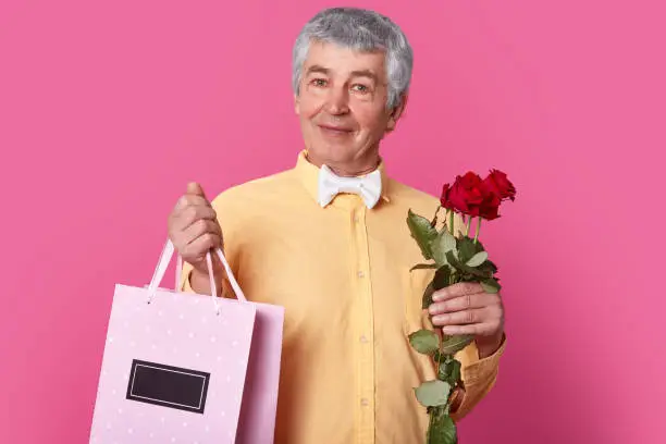 Handsome elegant man wears formal outfit, holds giftbag and bouquet of flowers, going to make present to his wife, being in high spirit, isolated over pink background. People, pension, celebration