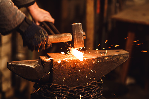 The blacksmith manually forging the red-hot metal on the anvil in smithy with spark fireworks