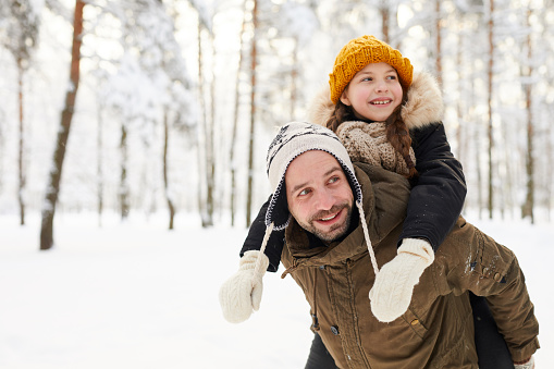 Portrait of happy little girl riding on fathers back in winter forest having fun, copy space