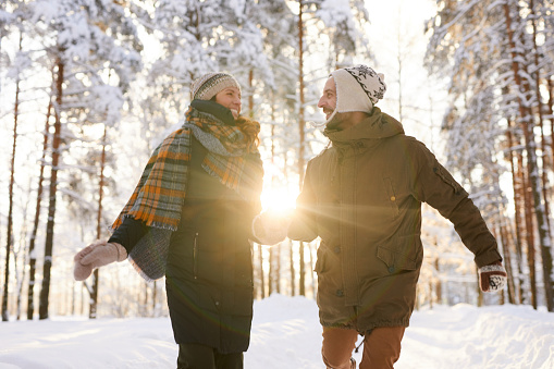 Portrait of playful couple running towards camera in winter forest and smiling happily lit by  sunlight, copy space
