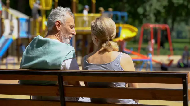 Elderly man and woman communicating on bench in park, watching grandchildren