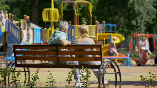 Senior couple sitting on bench near playground, watching grandchildren playing