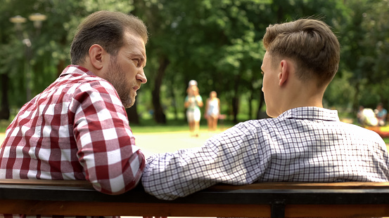Bearded father communicating with teenage son, sitting on bench in park, trust