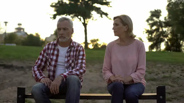 Sorrowful senior couple sitting on bench, wife looking at depressed husband