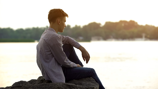 Melancholic male teen sitting alone on stone near river and thinking about life
