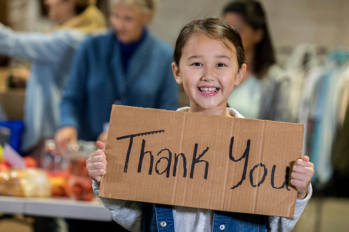 Smiling mixed race elementary age girl volunteers with her family in a local food bank. The girl is holding a 'Thank You' sign while smiling at the camera.