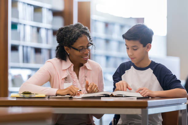 Tutor working with middle school student Encouraging female tutor works with preteen boy in the campus library. They are looking at a textbook. tutor stock pictures, royalty-free photos & images