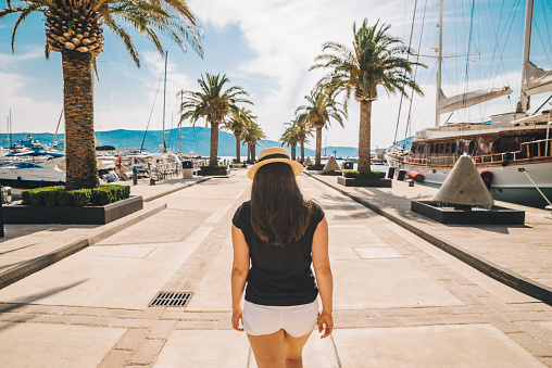 woman walk by sunny pier with palms. mountains and yachts on background