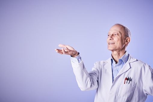 A mature man in a lab coat - possibly a scientist or medical professional - smiles as he gestures, holding aloft space for your message or product.