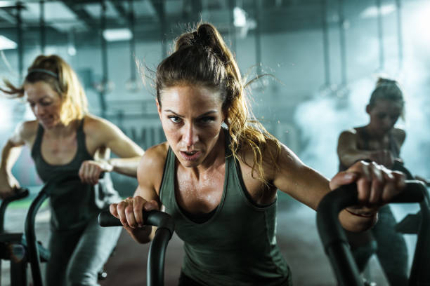 Determined athletic woman during exercise class on bike in a gym. Young female athlete having cross training on bikes with her friends in a gym and looking at camera. spin bike stock pictures, royalty-free photos & images