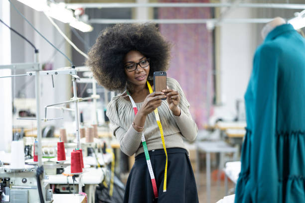 Designer de moda africana criando novos produtos na fábrica e usando telefone inteligente - foto de acervo