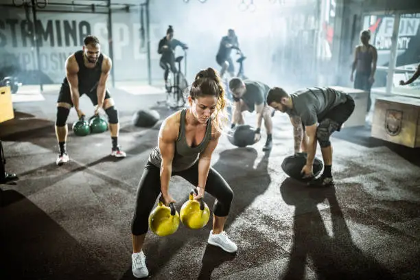 Large group of athletic people having training in a gym. Focus is on woman with kettle bells.