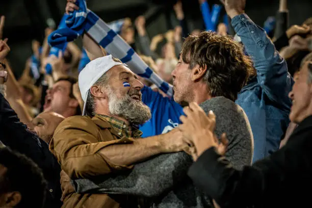 Group of football fans cheering and embracing each other while watching match in stadium.