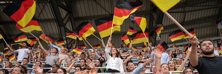 Fans holding German Flags and cheering while watching match in stadium.