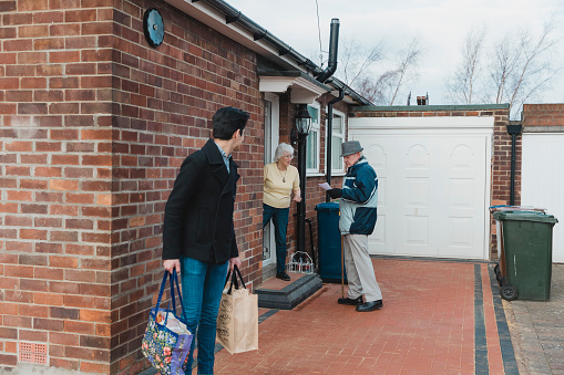 Teenager and his grandfather are leaving to go shopping together. The grandmother is standing in the doorway of their home and is giving her husband a shopping list.