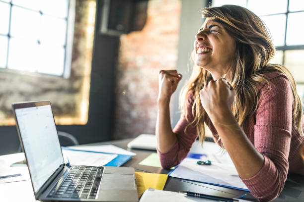 Cheerful woman received a great e-mail over computer. Cheerful casual businesswoman celebrating her achievement while using laptop in the office. punching the air stock pictures, royalty-free photos & images