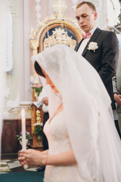 istock Groom looking at his bride under veil while she is praying with candle light. Happy stylish wedding couple holding candles  during holy matrimony in church. 1130459749