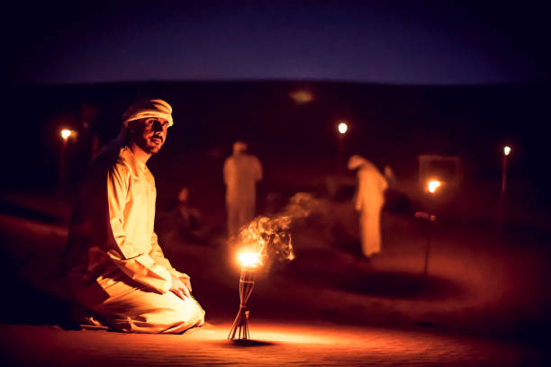 arab mean seated on the sand dunes near an oil lamp at a camp site - god of fire stock-fotos und bilder
